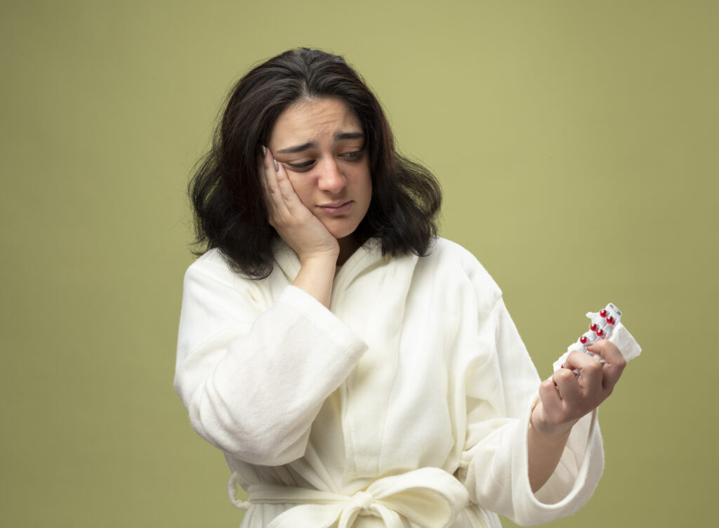 anxious young caucasian ill girl wearing robe holding napkin pack pills looking them isolated olive green background with copy space