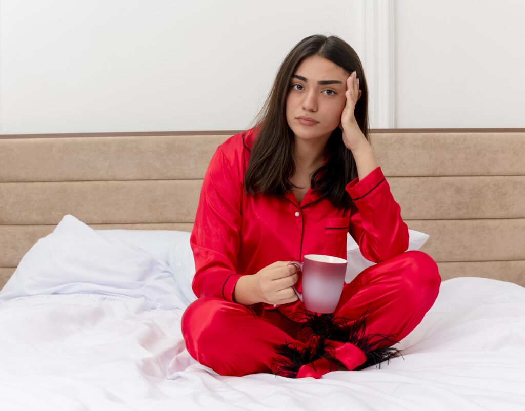 young beautiful woman red pajamas sitting bed with cup coffee looking camera being confused bedroom interior light background